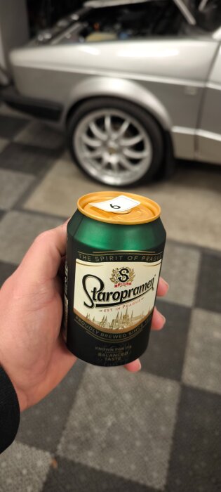 En hand håller en burk med Staropramen öl med en nummerlapp på locket, en bil syns i bakgrunden.