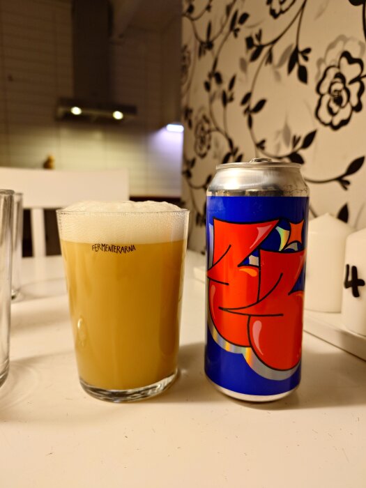 Glas med skummande dryck bredvid en färgglad dryckesburk med graffiti-liknande design på ett bord, insida ett rum med mönstrad tapet.