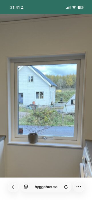 Vy genom ett fönster med vit karm, utsikt mot ett vitt hus med skog i bakgrunden, en kruka på fönsterbrädan.