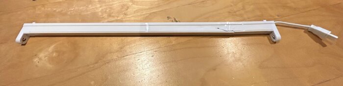 Trasig köksarmatur i vit plast, sprucken på mitten, liggande på ett trägolv med synlig elkabel på ena sidan.