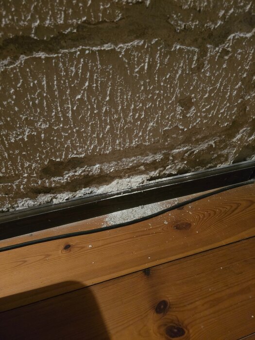 Vägg med sprucken puts ovanför ett trägolv, där bitar av vit puts har fallit ner.