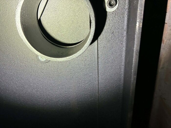 Rund öppning i en kamin, delvis täckt av en metallkant och en sten bakom. Hålet verkar inte fullt öppet trots att metallskenan dras bort.