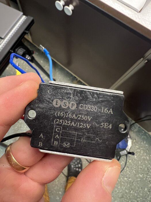 En person håller en svart elektrisk komponent märkt med specifikationerna 16A/250V och 25A/125V, med synliga anslutningskablar.