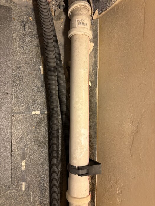 Vit avloppsrör och svart korrugerad slang fästa mot en vägg i ett rum under renovering.
