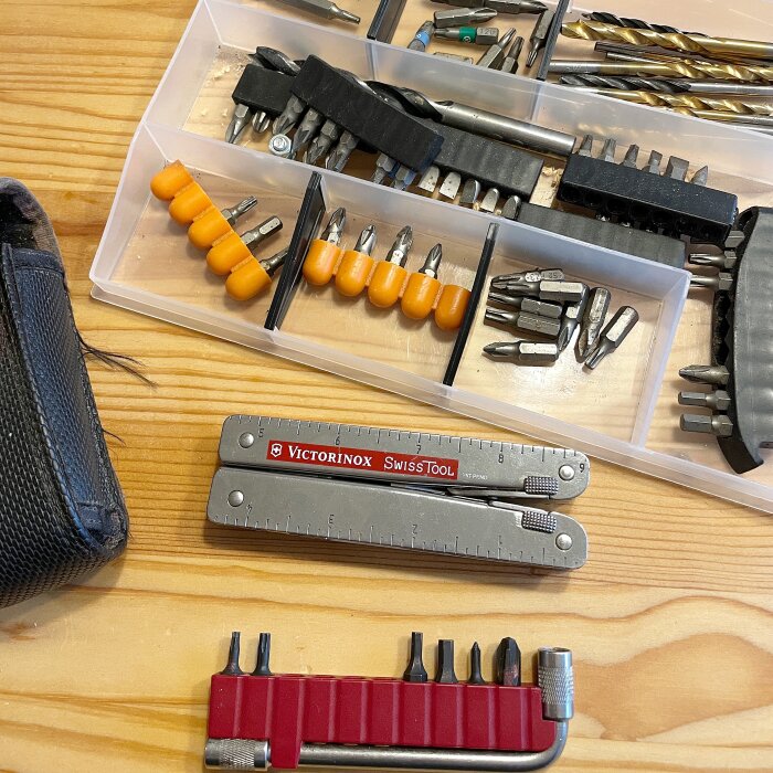 Bitsats och Victorinox SwissTool Plus på ett träbord. Verktygslåda med olika bits och borrar synlig.