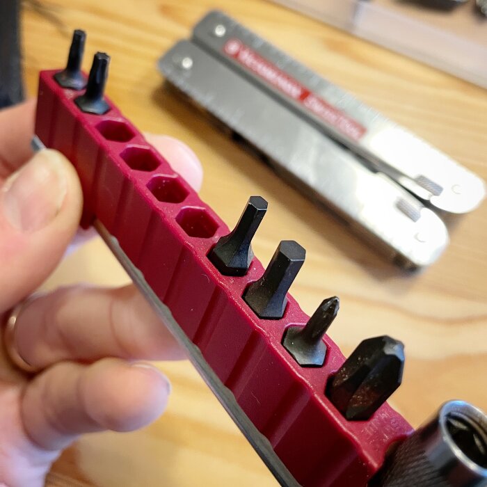 Röd bitsats med olika bitar, inkl. PH0 och PH3, hålls i handen; Victorinox SwissTool Plus syns suddigt i bakgrunden.