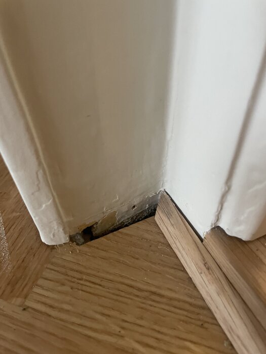 Närbild av parkettgolv intill dörrkarm, där golvet inte når helt under karmen. Kvartsstav synlig vid skarven, med ett mindre mellanrum.