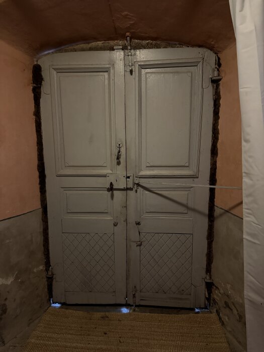 Gammaldags grå dubbeldörr i en tegelvägg, med litet ljus som sipprar igenom under dörren. Dörrarna har enkel, rektangulär paneldesign.