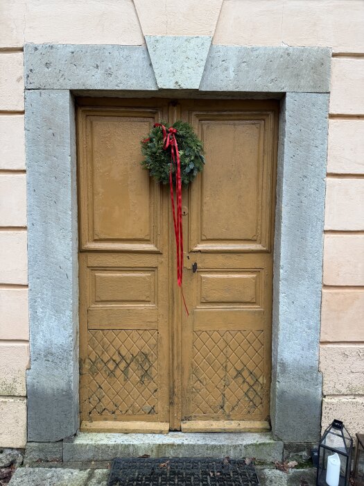 Gammal, brun, dubbel trädörr med en julkrans av granris och röd rosett hängande i mitten. Ljuslykta med vit blockljus bredvid dörren.