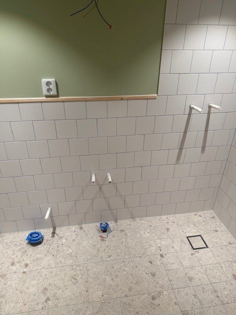 Pågående installation av badrum i attefallshus med vita kakelväggar och synliga PEX-slangar för vatten. Golvet är ljust och fläckat.