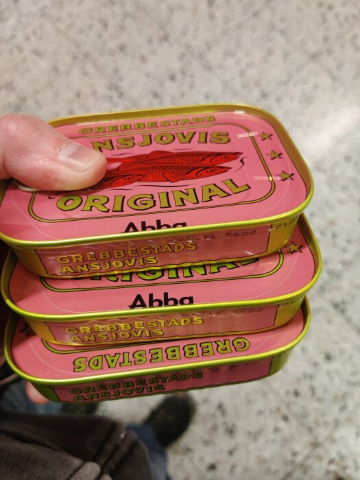 Hand som håller i tre burkar av Grebbestads ansjovis med rosa etiketter och texten "Original".