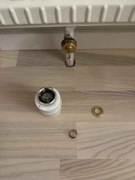 Termostatdelar och adaptrar bredvid en vattenburen radiator, med en ventil och metallring på golvet.