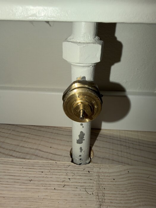 Närbild på en vattenburen radiatorventil med en kopplad termostat och adapterring, monterad på ett vitmålat rör mot golvet.