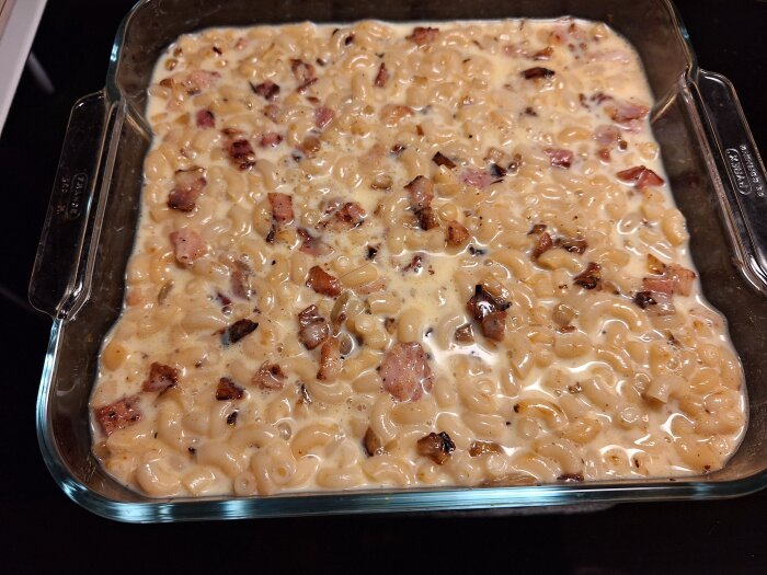 Makaronpudding i glassform med synliga bitar av bacon färdig att sättas in i ugnen.