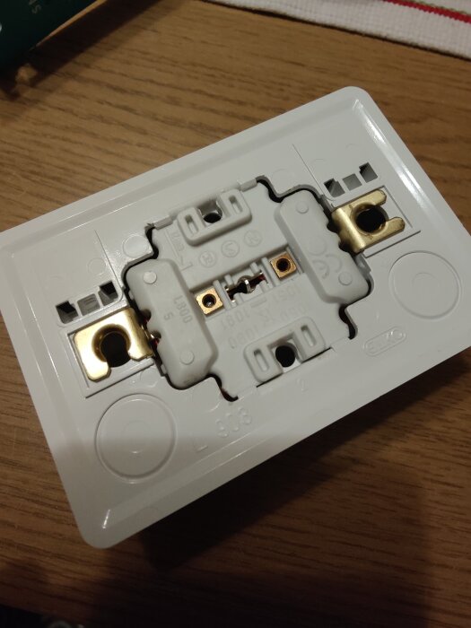 Vit elektrisk kopplingsdosa med synliga mässingsfästen och flera plastdetaljer, placerad på ett träbord.