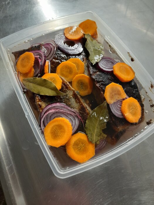 Inlagd fiskfilé med skivade morötter, rödlök och lagerblad i en plastbehållare på ett metallbord.