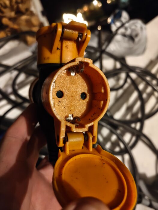 Ett orange trevägsuttag med synlig saltbeläggning inuti, hålls upp av en hand. Bakgrunden visar elektriska kablar.
