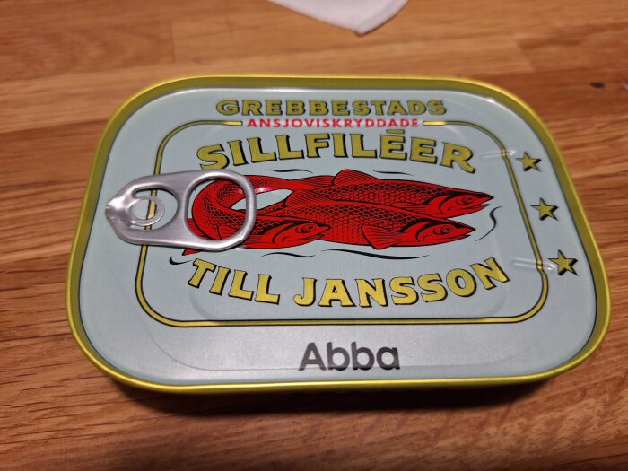 Konservburk med ansjoviskryddade sillfiléer från Grebbestad, text "Till Jansson" och Abba-logga, på en träyta.