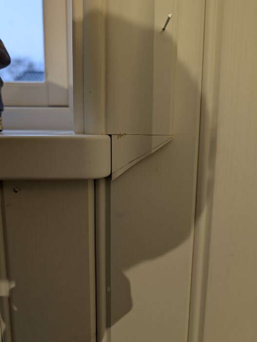 Hörn av fönsterkarm med temporärt träfoder monterat, fäst med synligt fäste, skuggor och bakgrund av fönster och väggpanel.