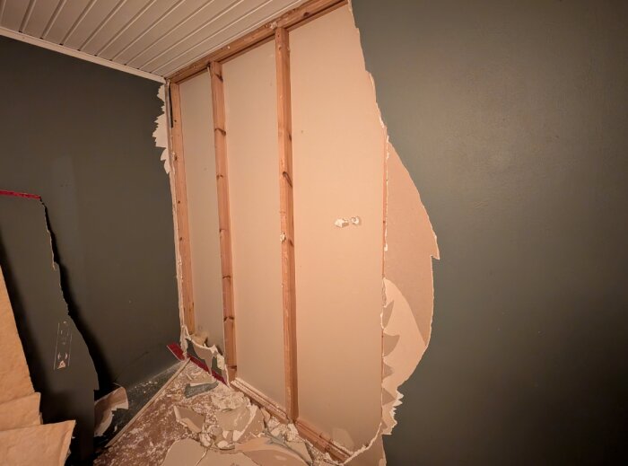 Riven gipsskiva visar träreglar i en innervägg under renovering, med gipsspill på golvet.