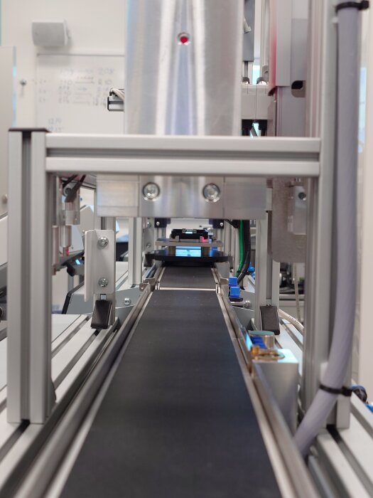 Industriell maskin med transportband i en fabriksmiljö, med synliga metallkomponenter och teknisk utrustning.