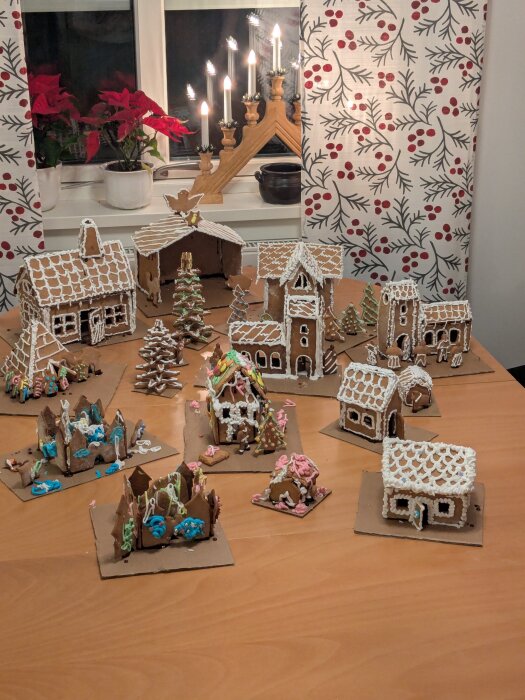 Familjens pepparkakshus, dekorerade med färgad glasyr, stående på ett bord framför ett fönster med ljusstake och julstjärnor.