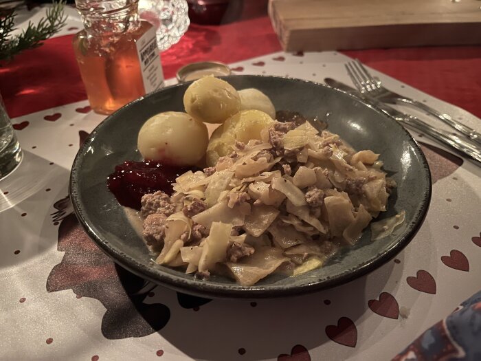 Tallrik med Kålmaja, potatis, lingonsylt och chiliäppelgelé, serverad på röd duk med hjärtmönster.