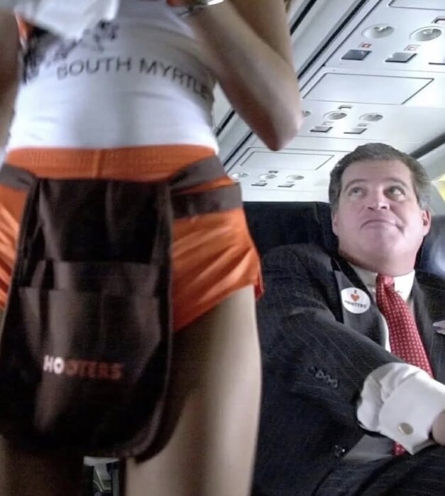 En man i kostym sitter i ett flygplanssäte och tittar uppåt. En person i Hooters-uniform står framför honom.
