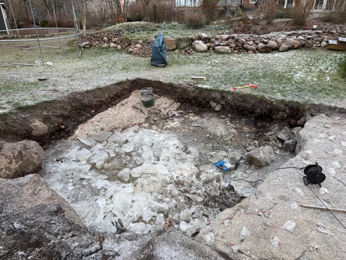 Uppgrävd mark fylld av krossad is, stenblock och ett verktyg i en trädgårdsmiljö, med arbetsredskap i bakgrunden.