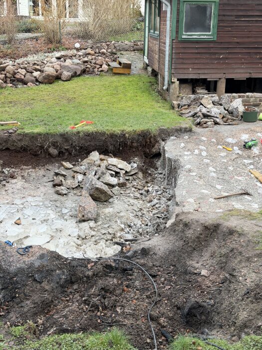 Bild på en utgrävd plats bredvid ett hus med brädfasad och grönt fönster, stenhögar och gräs intill. Visar arbetet med att bryta sten.