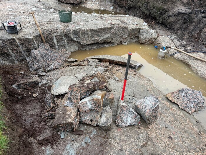 Sprucket berg med lösa stenblock, en yxa och en slägga, samt vattenpöl i bakgrunden med en pump i ett utomhusområde.
