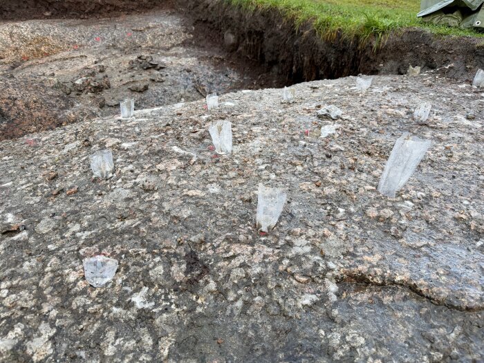 Borrhål i berget för spräckning med snigeldynamit. Plaststrumpor är insatta i hålen, omgivna av sprickor på en ojämn bergyta.