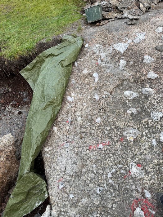 Berg med borrhål och sprickor, täckt delvis av en grön presenning. Stenbitar och en grön hink syns ovanpå berget.