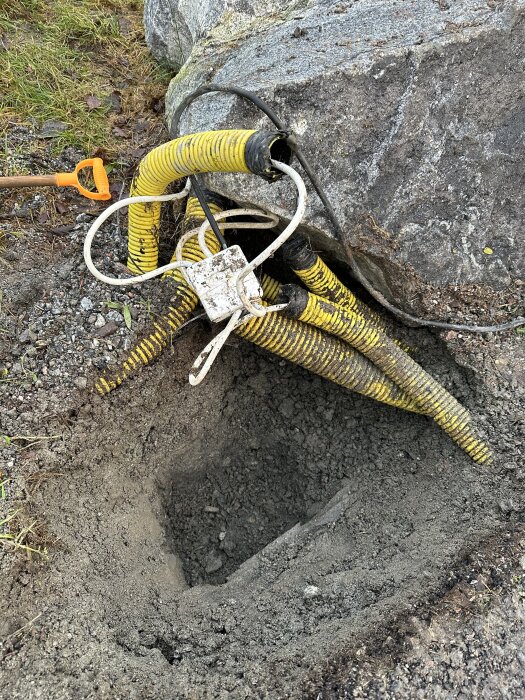 Gulrör och elkablar bredvid en stor sten med en oöppnad kopplingsbox fyllt med slime, omgiven av grävd jord för fundament till laddstolpe.