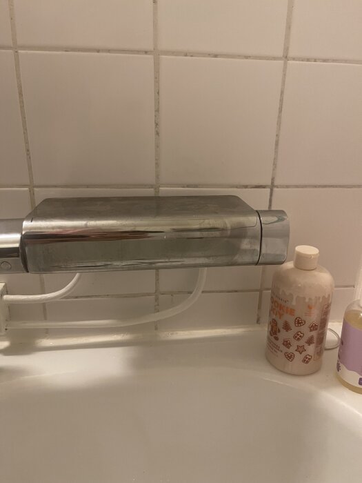 En kromfärgad dusch/badkarsblandare i en vitkaklad dusch med en pip som har lossnat, bredvid står två flaskor.