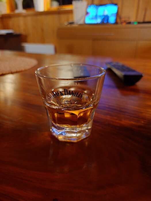 Whiskeyglas med Jack Daniel's-logotyp på ett träbord, TV syns suddigt i bakgrunden.