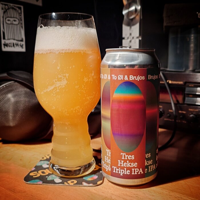 En glas fylld med ljus IPA och en burk med etiketten "Tres Hekse" på ett bord, bredvid ljudutrustning.