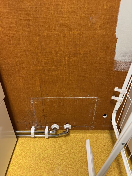 Rörinstallation på en orange vägg med två rör och vita fästen. Ett hål syns på väggens högra sida och en del av ett torkställ. Golvet är gult.