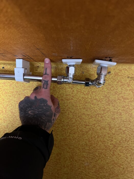 En tatuerad hand pekar mot rör under diskbänk med felaktig installation på orange vägg.