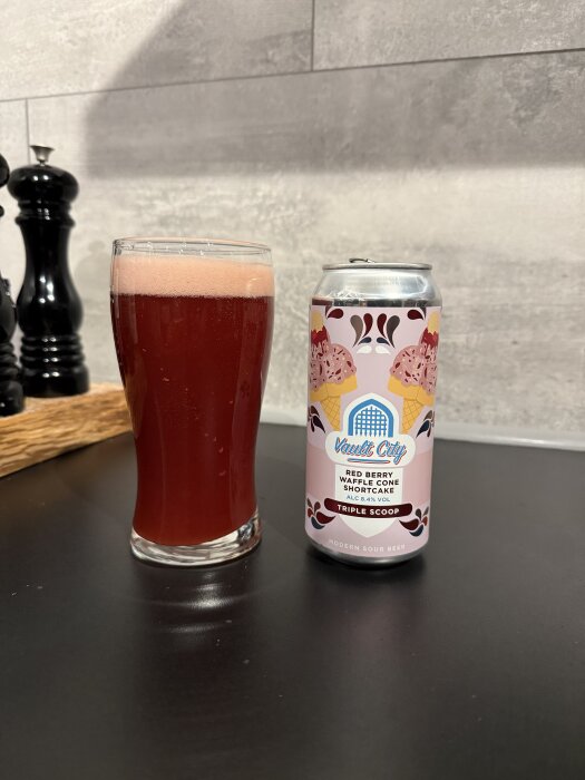 Ett glas röd dryck med skum bredvid en burk "Vault City Red Berry Waffle Cone Shortcake" modern sour beer på ett bord.