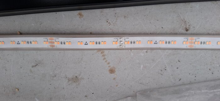 LED-strips med synliga dioder, lödningar och märkningar som visar 24V anslutningar, liggande på en betongyta.