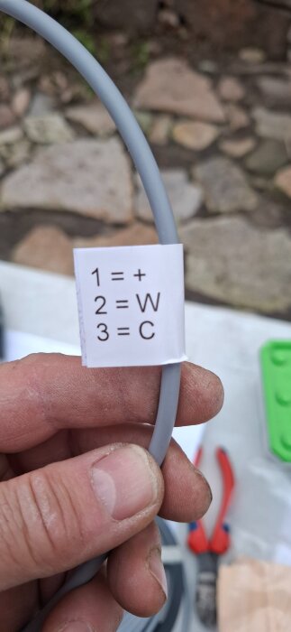 En hand håller i en tre-ledad anslutningskabel med vit etikett som visar anslutningar: 1=+, 2=W, 3=C. Bakgrunden visar stenar och verktyg.