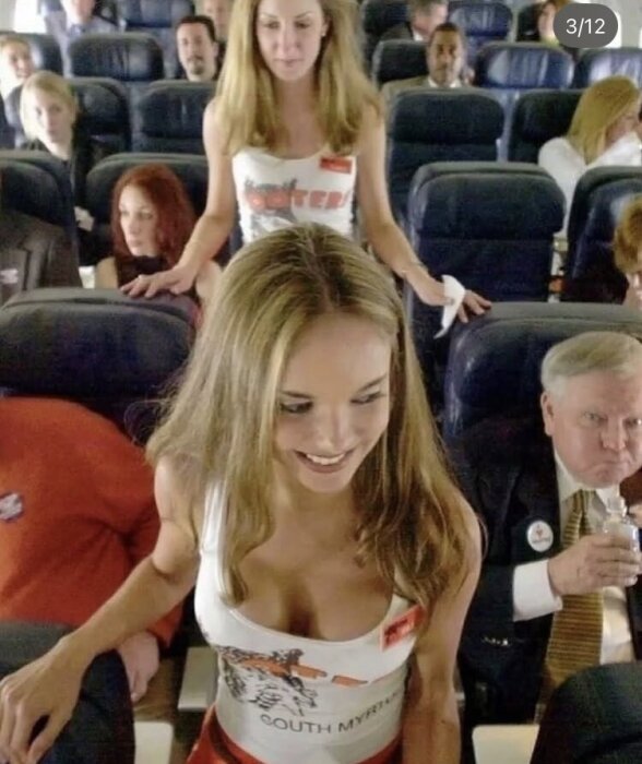 Människor sitter i flygplansstolar, två kvinnor i Hooters-linnen står i gången.