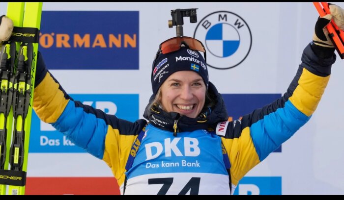Kvinna i gul och blå jacka med nummerlapp håller skidor och firar seger på en podie.