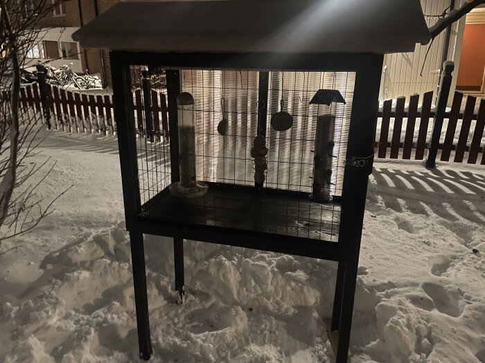 Pippimatstation av kompostgaller och spillvirke, belägen i en snötäckt trädgård, med fågelmatare och skyddande tannerplast på baksidan.