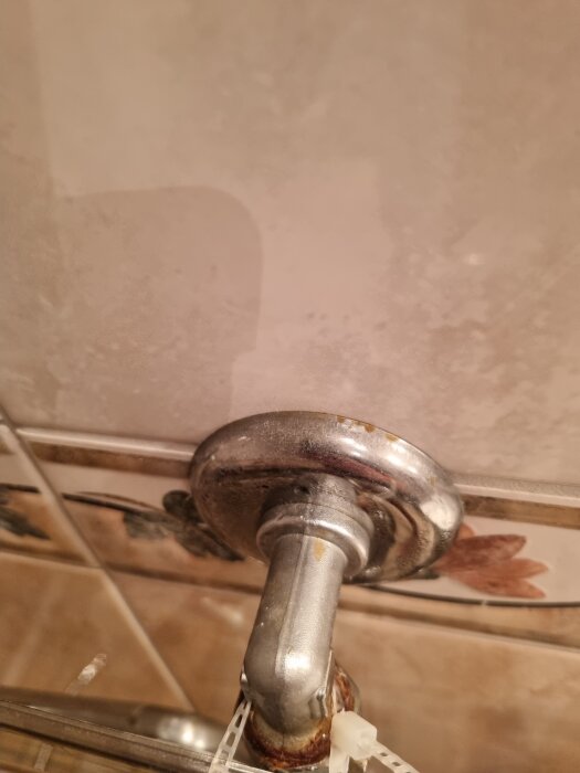 En närbild av en metallventil med rostfläckar mot en kakelvägg i ett badrum utan duschkabin.