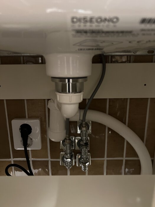 Vattenrör och röranslutningar under ett handfat, med synliga kopplingar och ett eluttag.