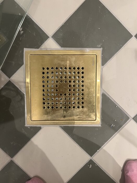 Golvbrunn i mässing omgiven av kakelplattor, vissa med synliga ojämnheter och smuts, vilket reflekterar dåligt utfört kakelarbete.