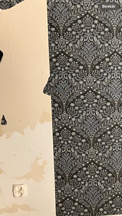 Riven tapet på en vägg, blottar vit yta och eluttag. Intill finns ett mönstrat tapetmönster med snirklar och blommor i mörka färger.