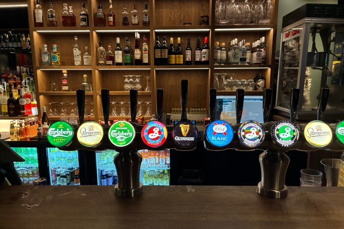 Ölkranar på en bardisk med olika ölsorter, inklusive Carlsberg, Staropramen och Guinness, och hyllor med flaskor i bakgrunden.